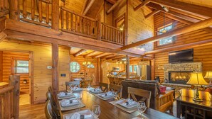 Ein Holzblockhaus-Innenraum mit Essbereich, gedecktem Tisch und Kamin aus Stein. Darüber ist ein Fernseher angebracht.