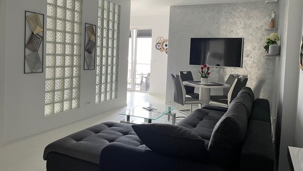 A modern living room with a sectional sofa, a glass coffee table, and a flat-screen TV mounted on the wall. - Westpark2 One-Bedroom Suite W5 Lakeview (Bucharest)