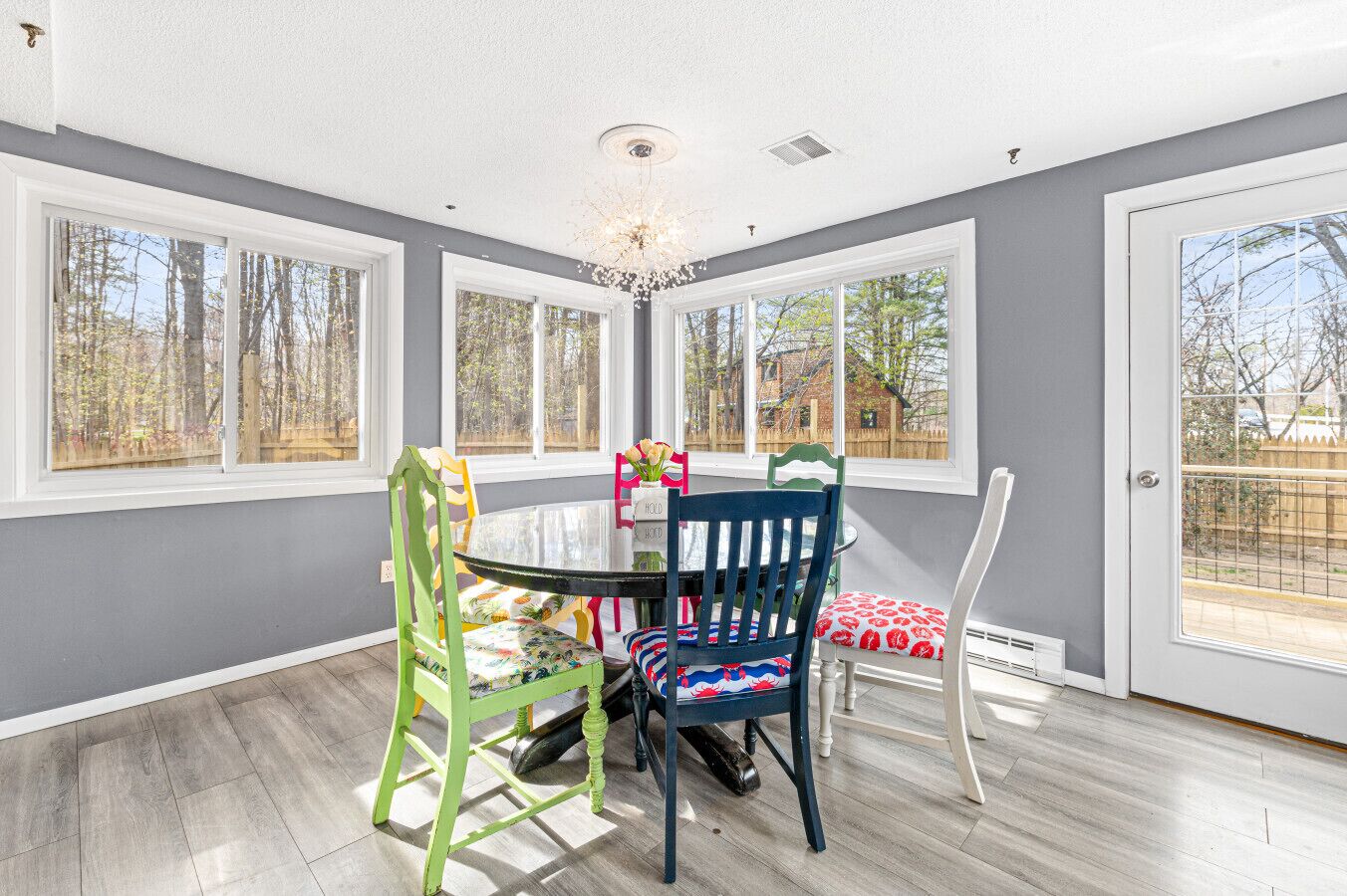 A dining area with a round table, colorful chairs, and large windows offering a view of trees and a fence.