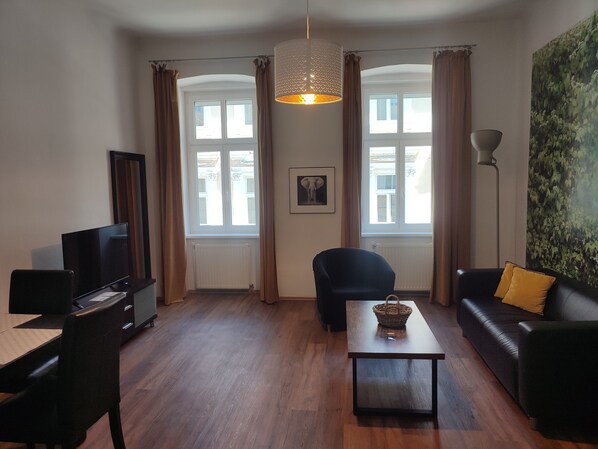 Uma sala de estar com televisão, sofá, poltrona, mesa de centro e uma janela grande com cortinas.