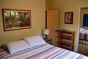 Un dormitorio con cama, un cuadro, una puerta, una lámpara y una mesita.