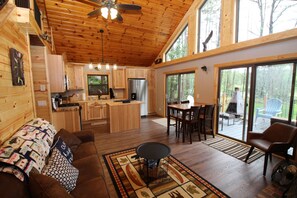 A spacious cabin interior with a wooden ceiling, a dining area, a kitchen, and a large sliding glass door leading to a deck. - Waterfront Home On Beautiful Lower Post Lake (Elcho)