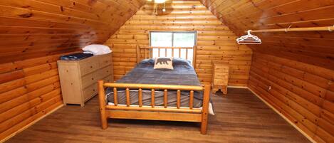 A small, cozy attic bedroom with wooden walls and a sloped ceiling. It features a single bed with a wooden headboard, a dresser, and a window.