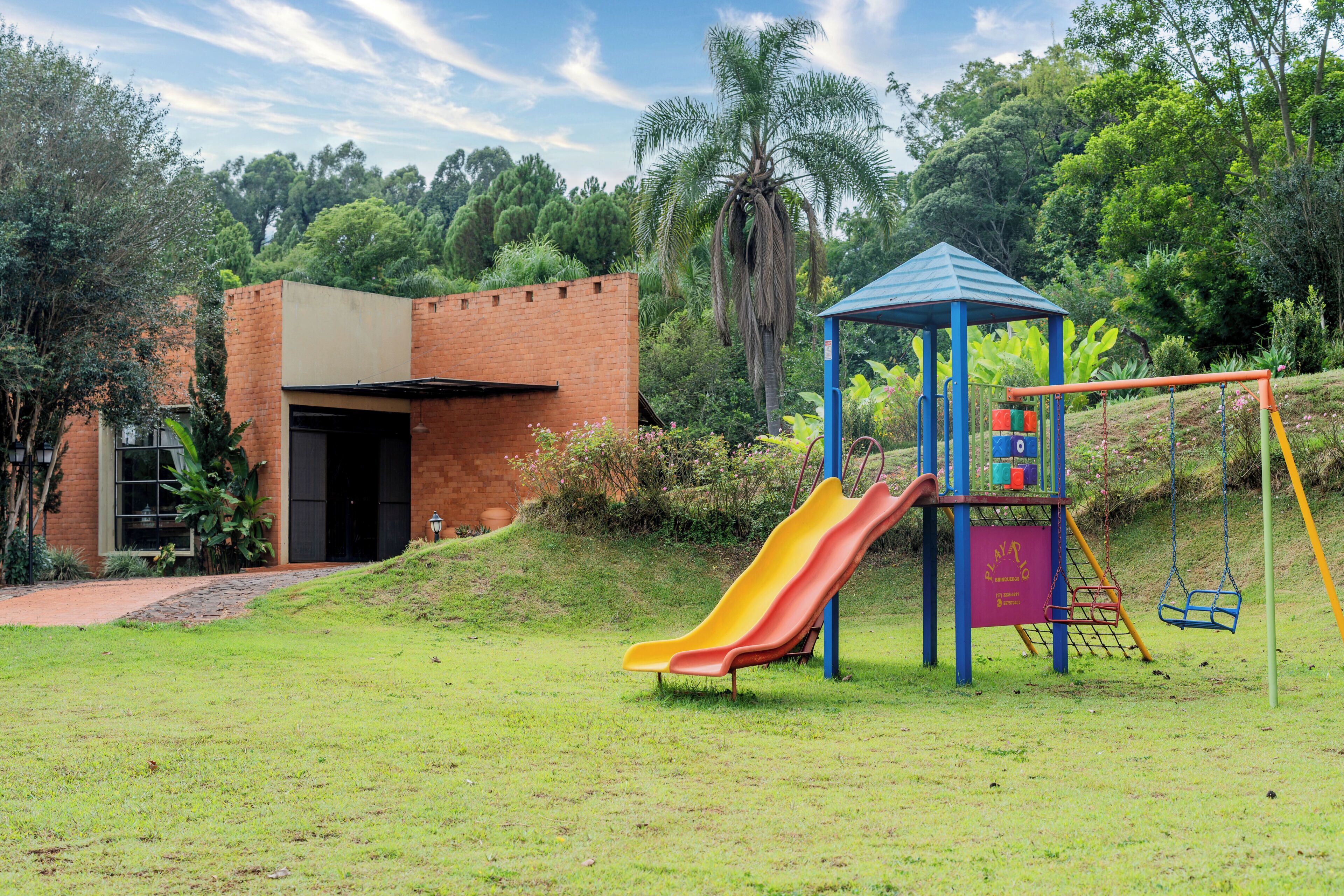 Um parquinho com escorrega e balanços, um prédio de tijolos com uma porta, e uma palmeira ao fundo.