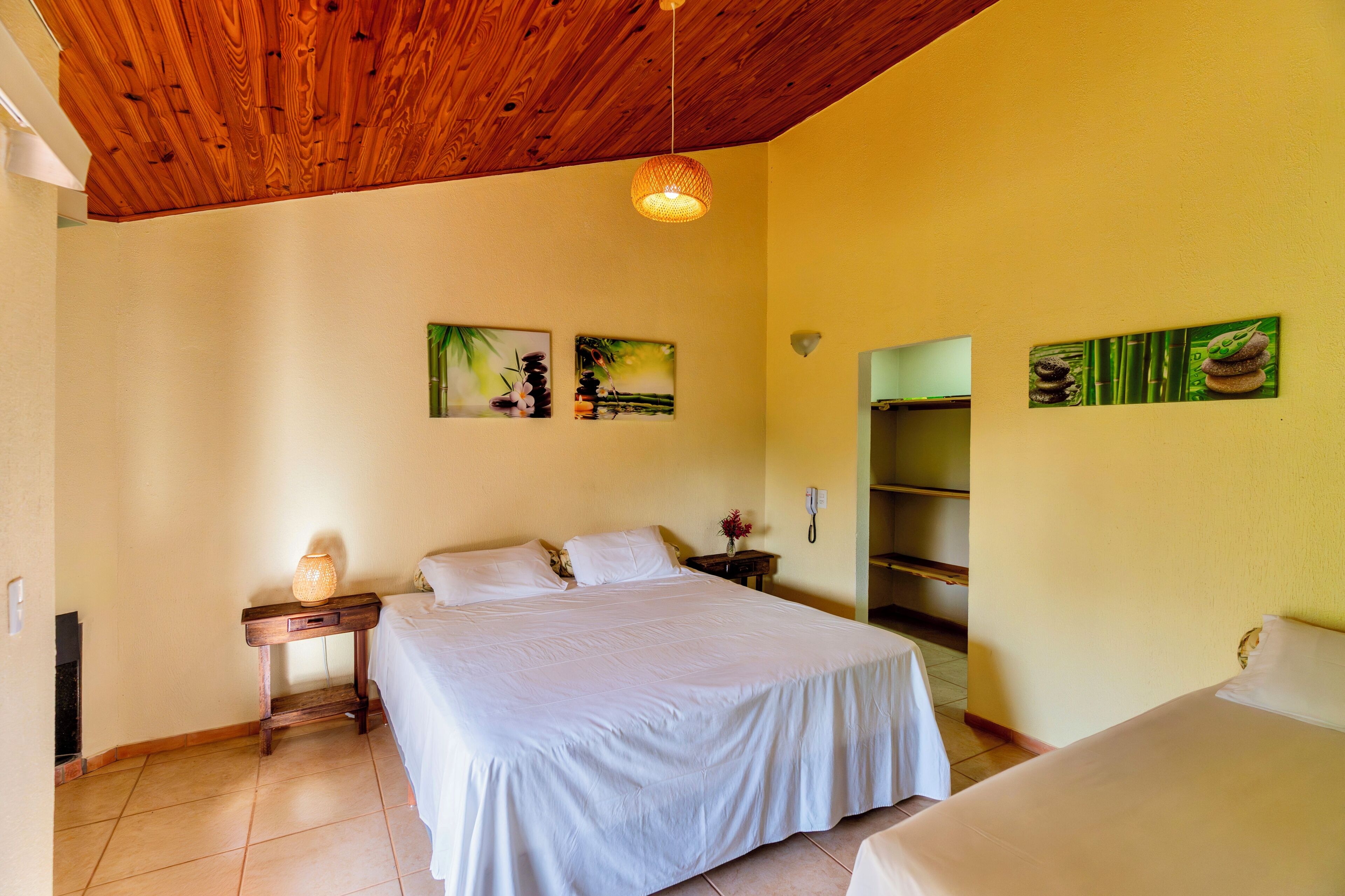 Un dormitorio con techo de madera, una cama con ropa blanca, una mesita de noche con lámpara y obras de arte en las paredes.