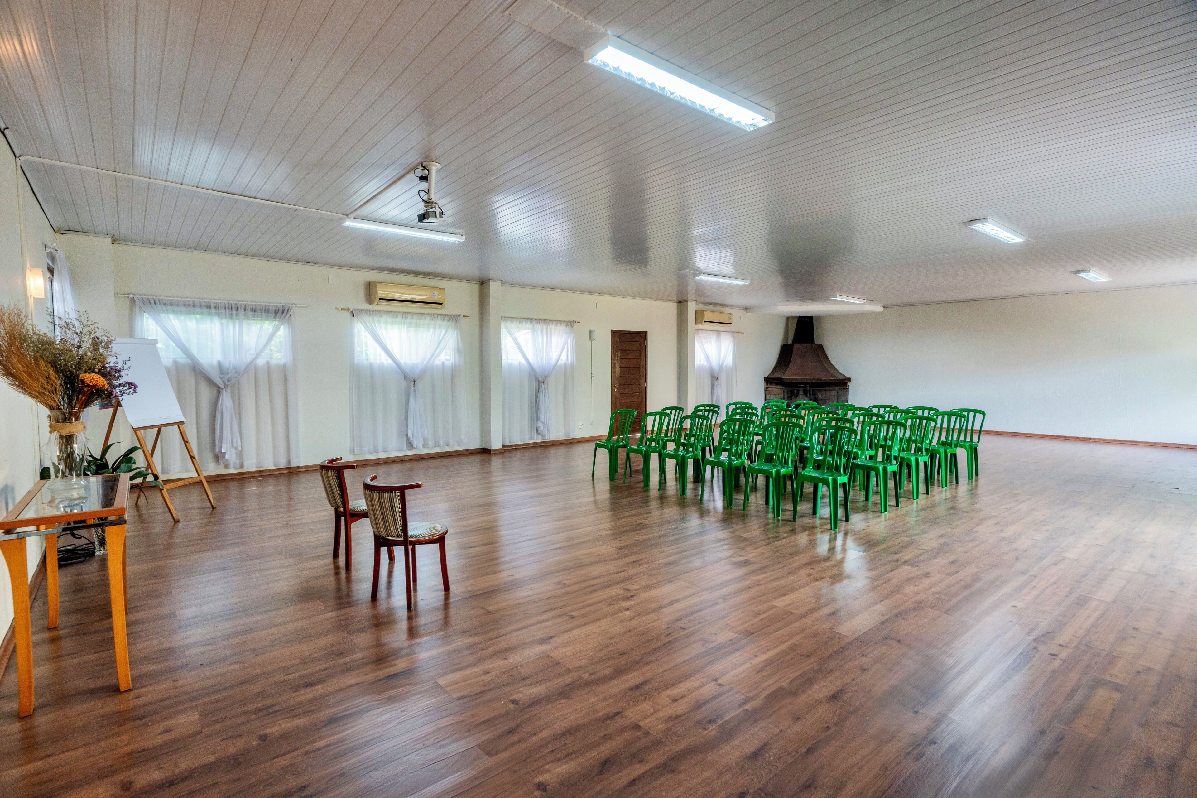 A spacious room with wooden flooring, a large fireplace, and green chairs arranged for a meeting.