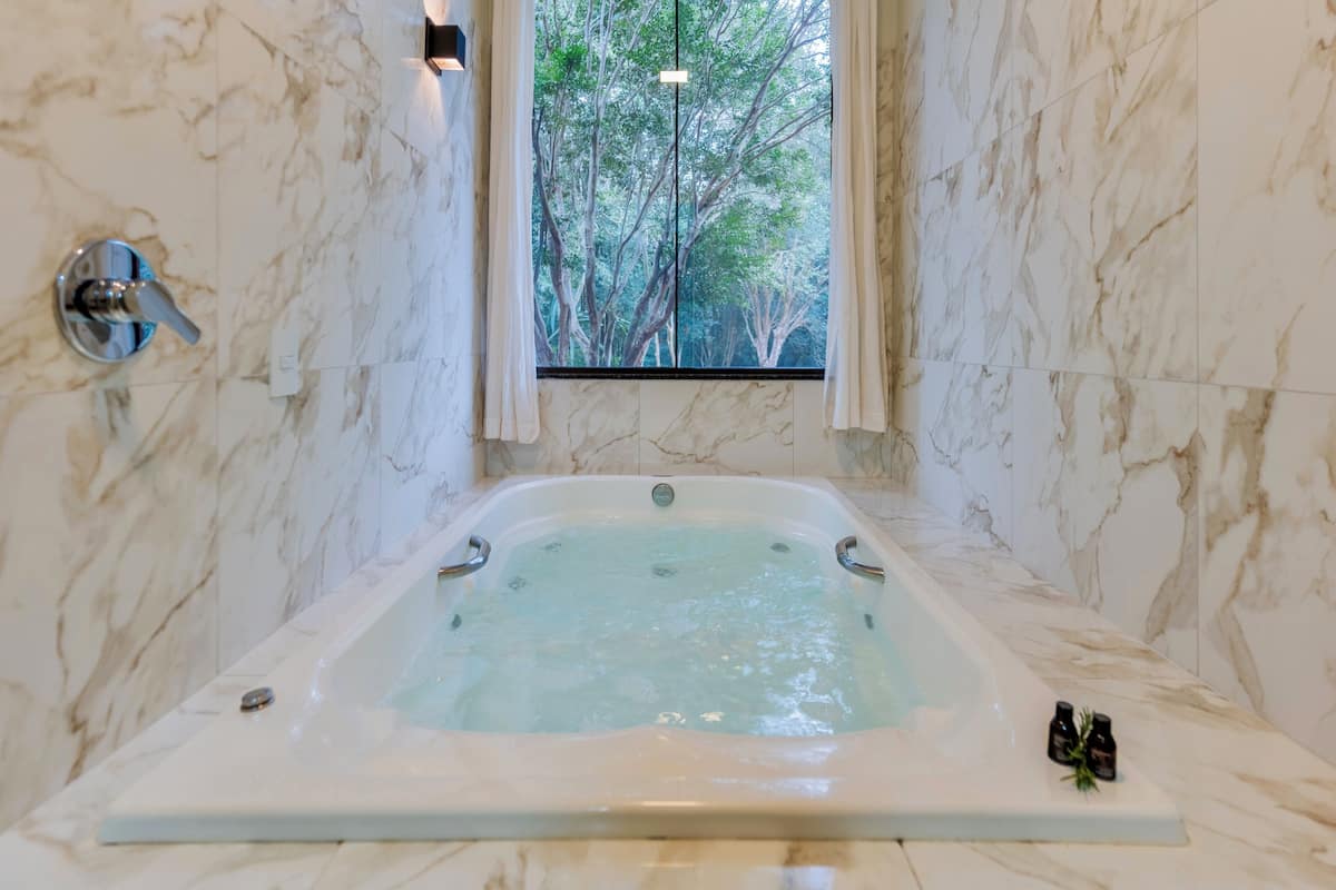 A marble bathroom with a bathtub, a window with a view of trees, and a small shelf with two bottles.