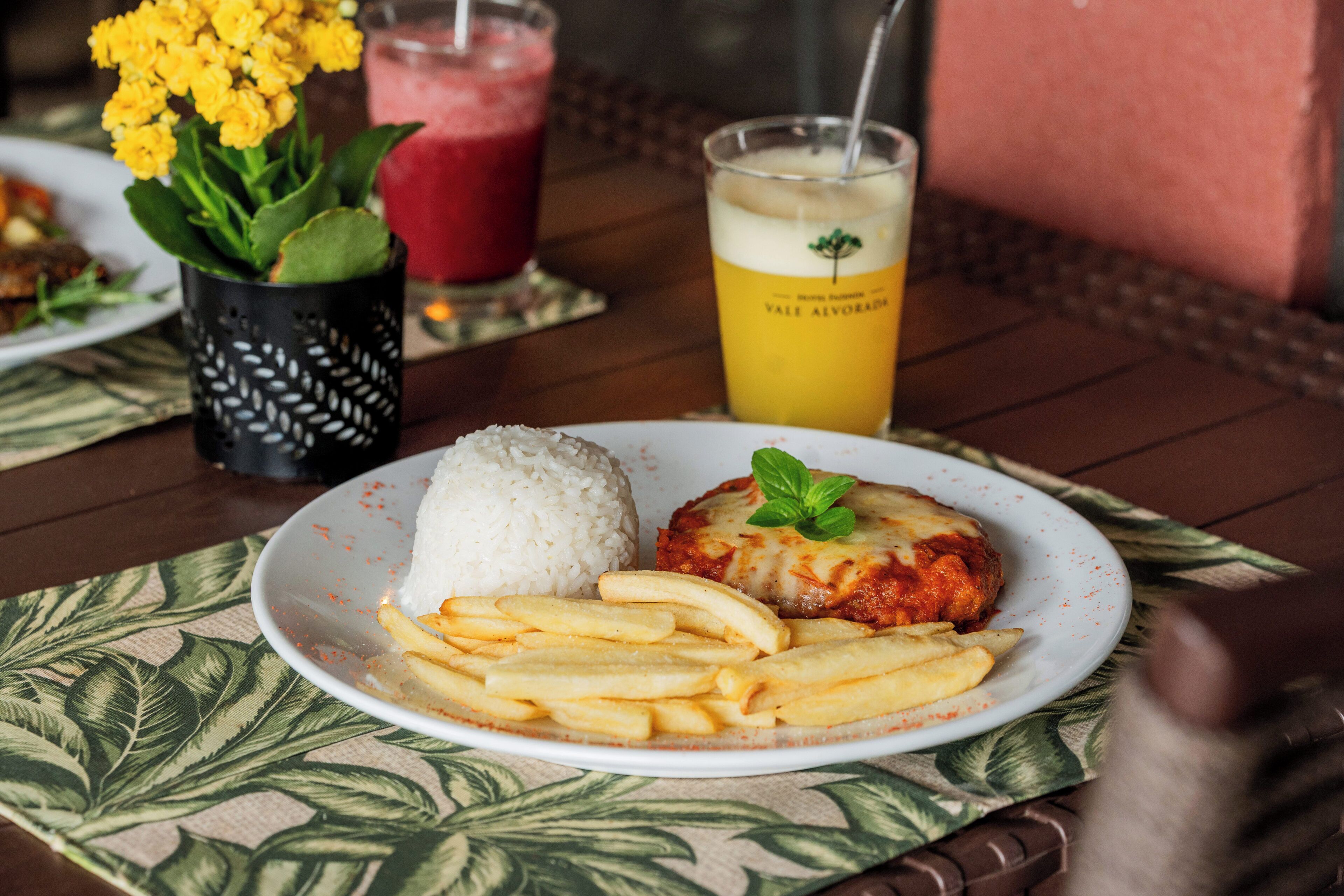 A plate with rice, a meat dish, and fries on a table with a drink and flowers in the background.