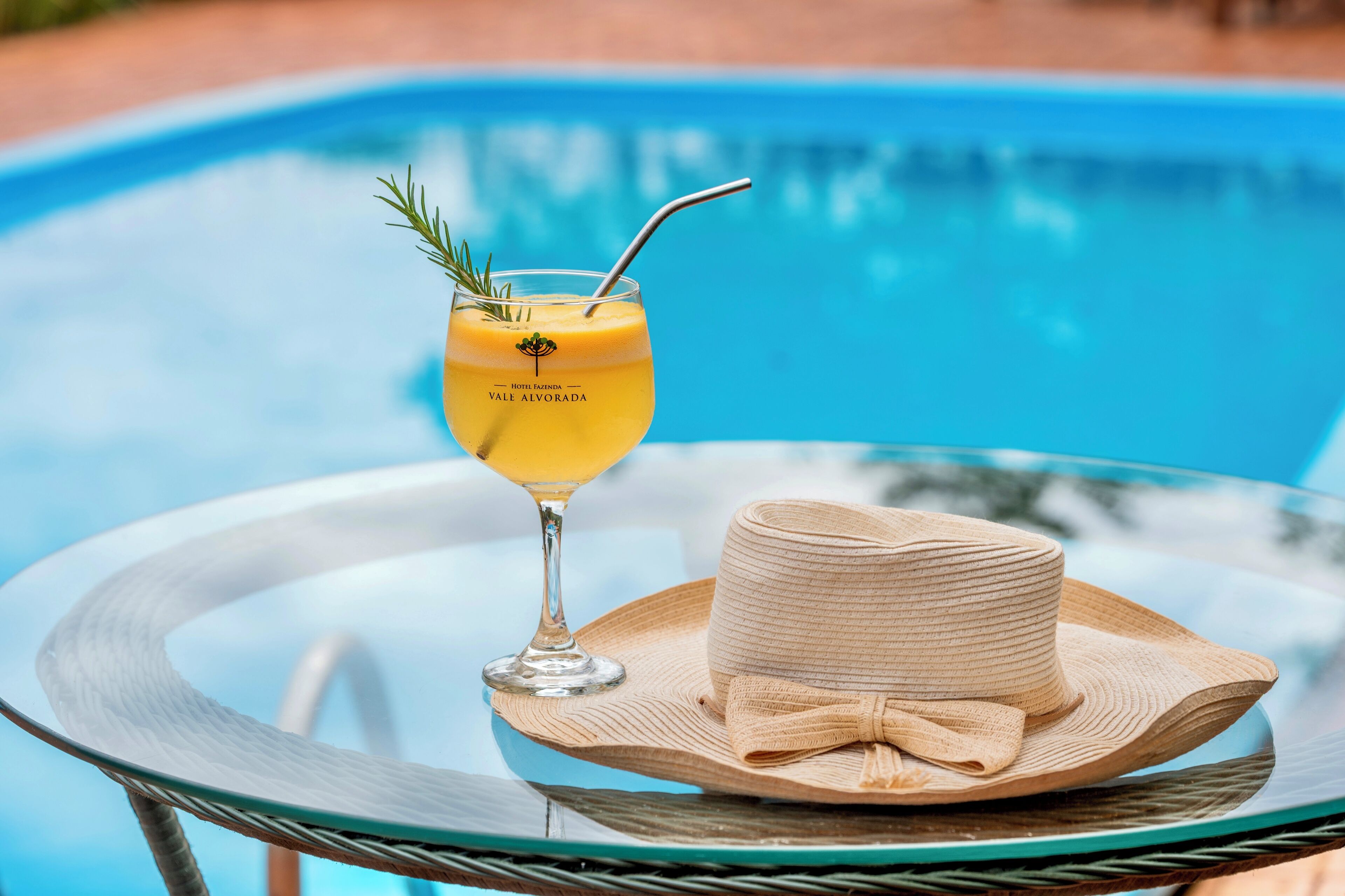 Un vaso de cóctel amarillo con pajita y ramita de romero sobre una mesa de cristal junto a una piscina.