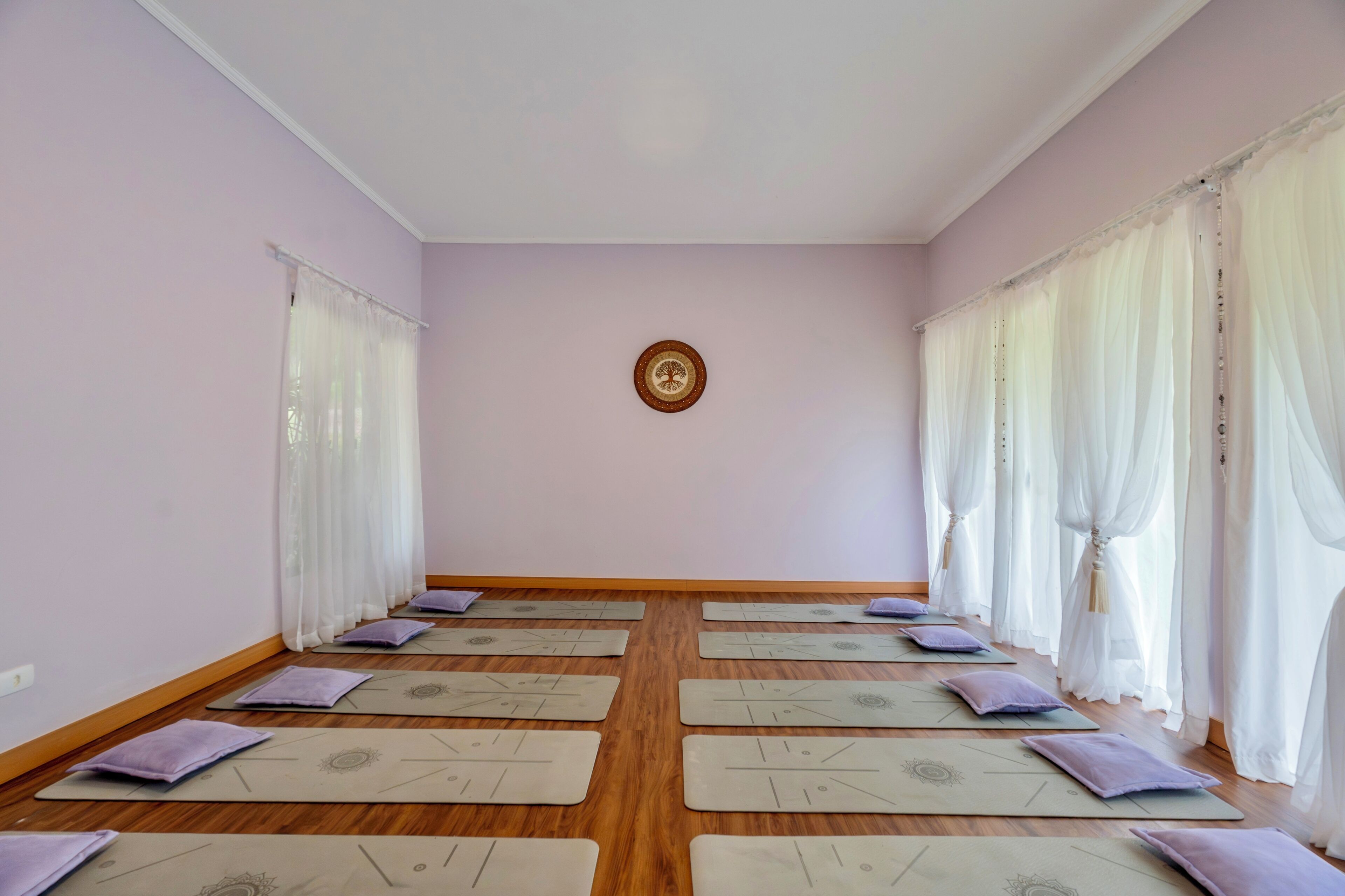 Um estúdio de yoga com tapetes dispostos em fileiras, piso de madeira e cortinas leves.