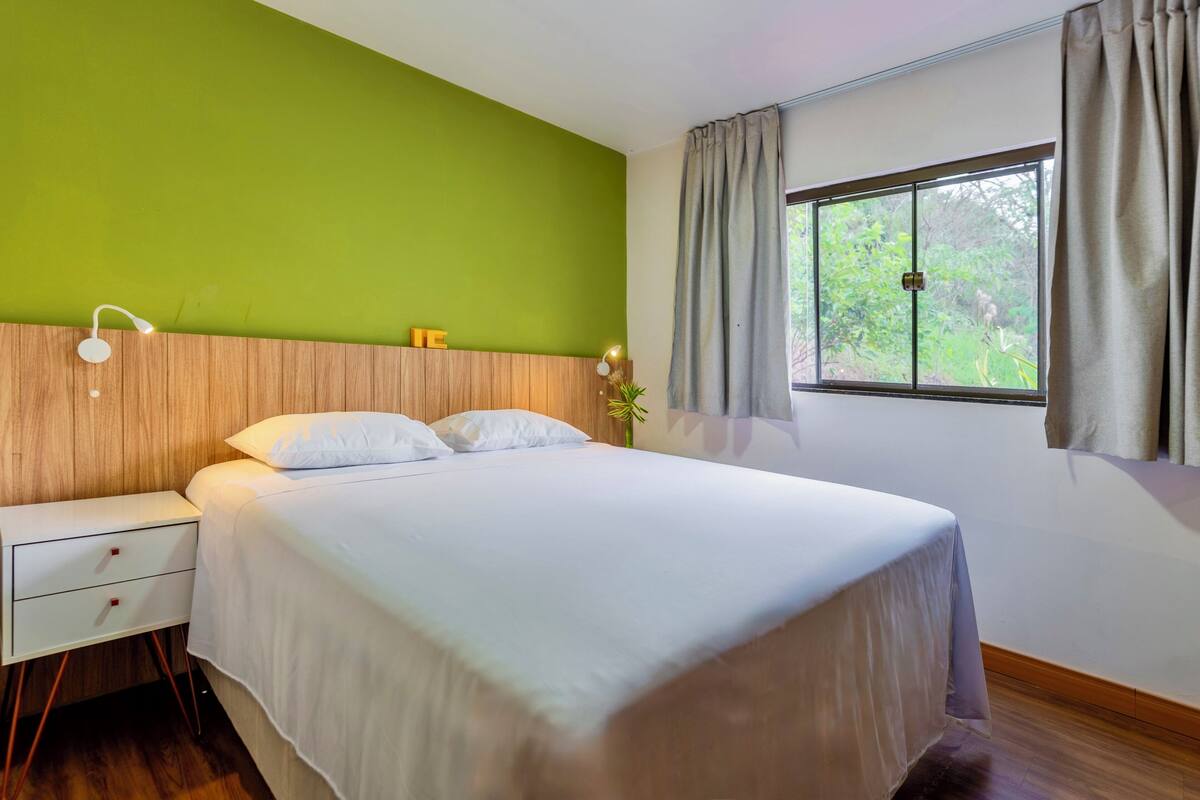 A neatly made bed with white linens, a wooden headboard, a bedside table with drawers, a window with curtains, and a green wall.