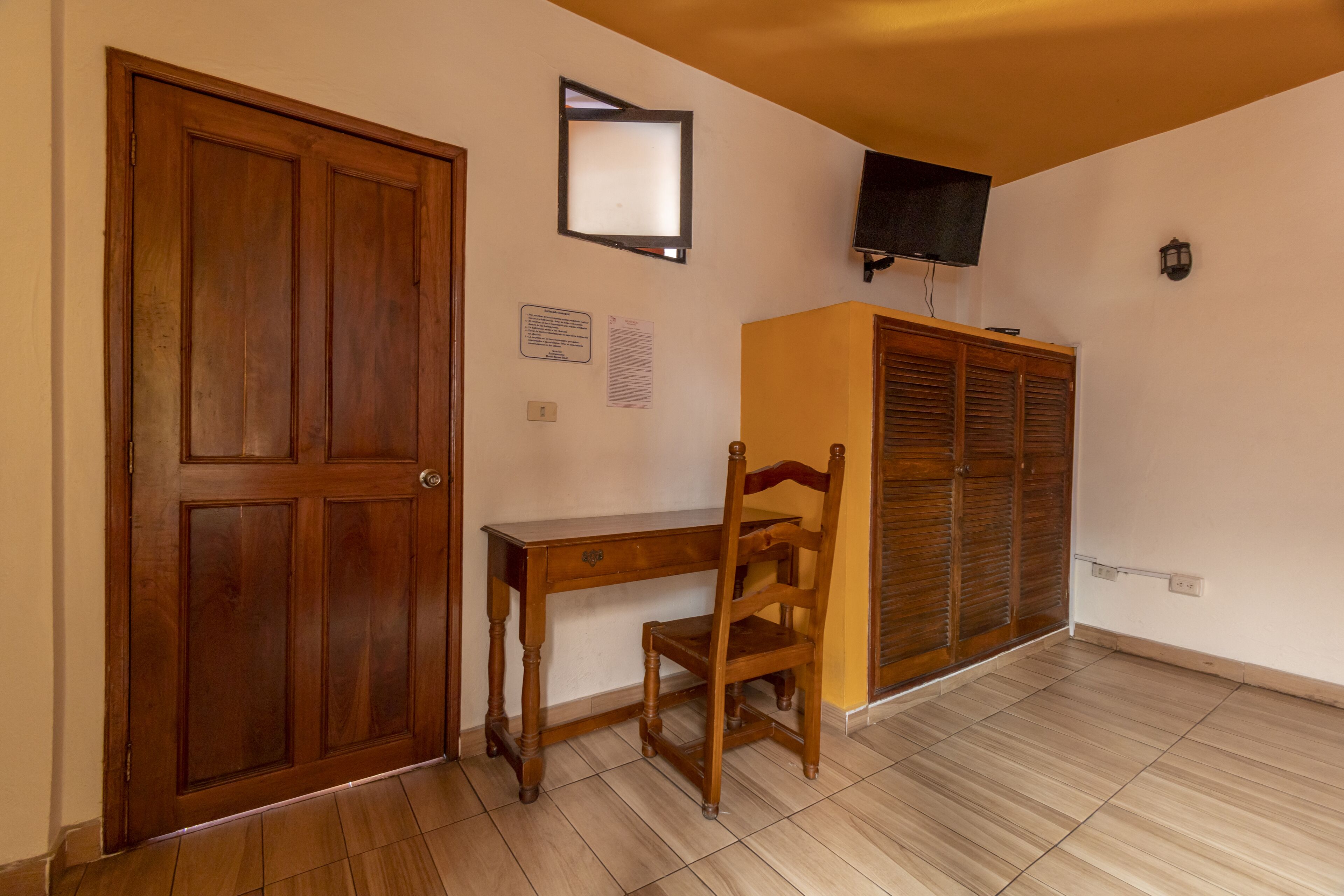 Una habitació amb una porta de fusta, un escriptori, una cadira, un televisor fixat a la paret i un armari.