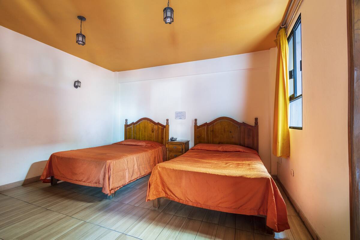 A room with two wooden beds, a small bedside table, a wall-mounted light, and a window with yellow curtains.