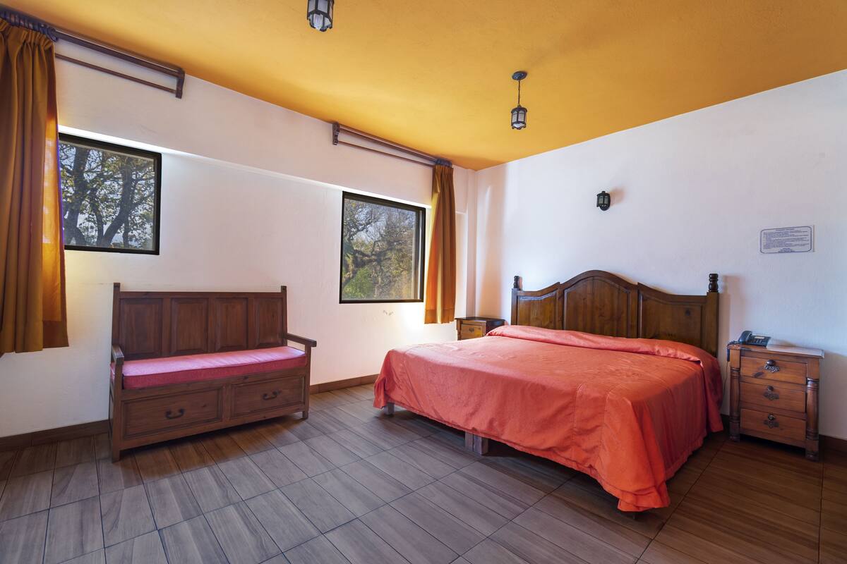 A bedroom with a wooden bed, a wooden bench, a nightstand, and two windows with curtains.