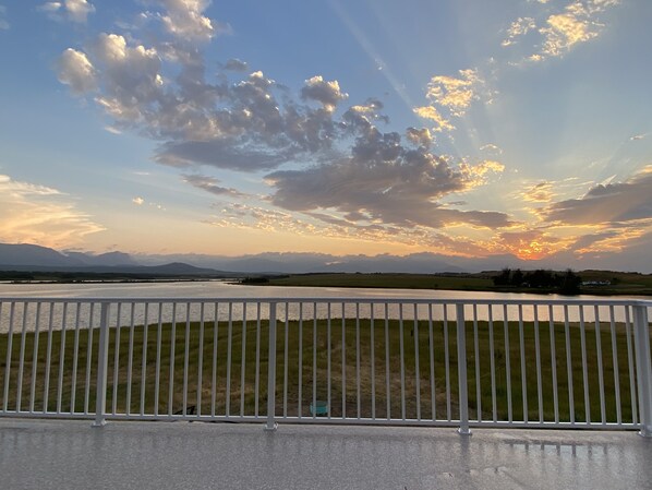 Une vue sur un lac, avec une chaîne de montagnes lointaine, un ciel dégagé parsemé de quelques nuages, et une rambarde métallique blanche au premier plan.