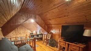 Ein gemütliches Dachzimmer mit Holzwänden und geneigter Decke. Es bietet ein bequemes Sofa, einen Fernseher und einen kleinen Tisch mit einer Lampe.