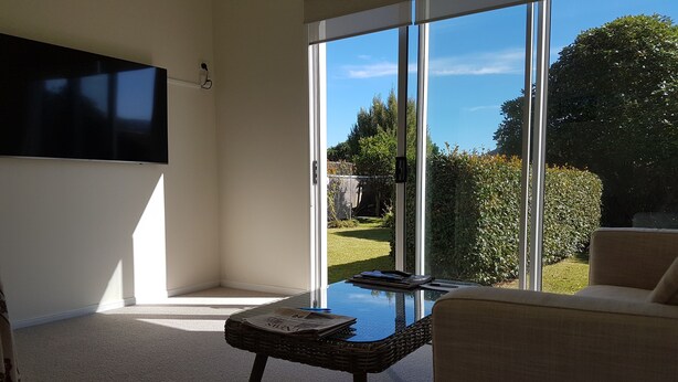 A modern living room with a large glass door leading to a garden, a flat-screen TV mounted on the wall, and a wicker table with newspapers.