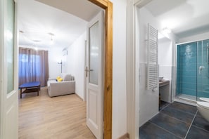 A room with a wooden floor, a white door, a white sofa with cushions, a small table, and a bathroom with a blue shower.