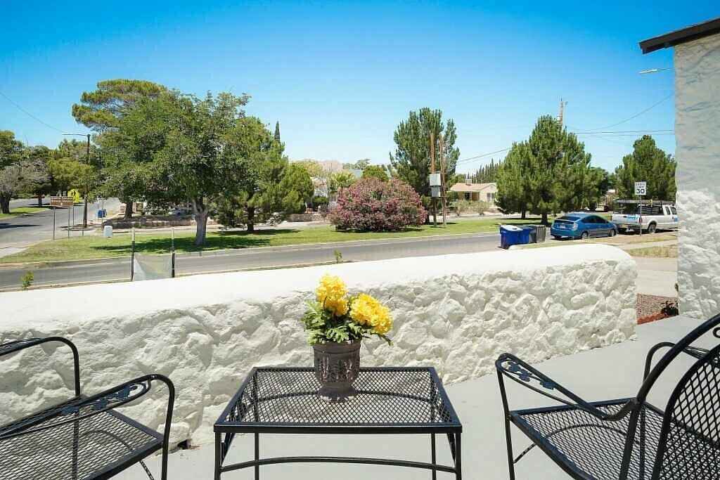 A patio with a table and chairs, a flower vase, and a view of a street with cars and trees.