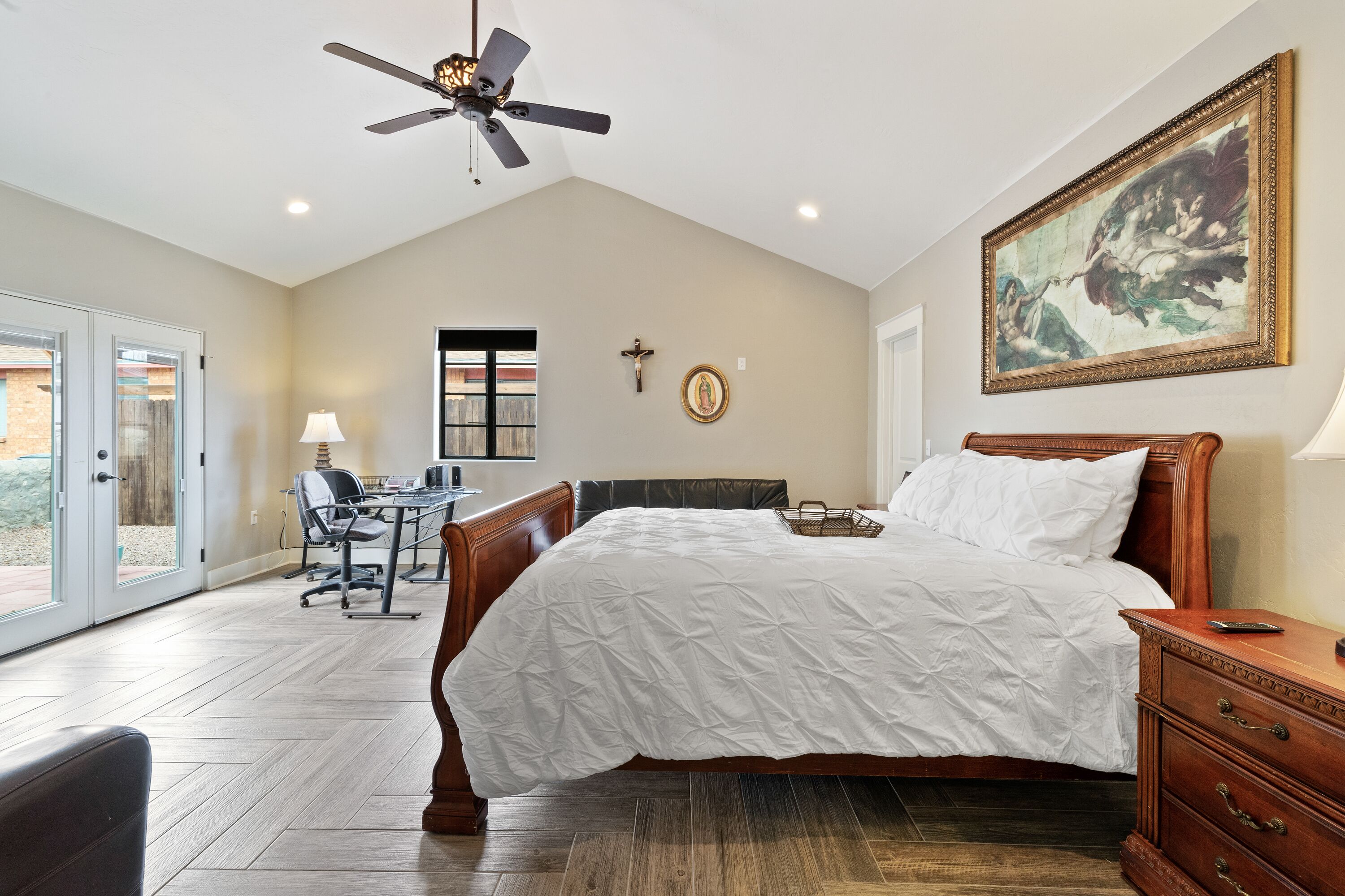 A bedroom with a bed, a desk, a chair, a ceiling fan, a painting, and a cross on the wall.