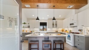 A modern kitchen with white cabinets, a central island with stools, pendant lighting, and a wooden ceiling.