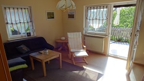Una sala de estar con un sofá, una mesita, una silla y un radiador. Hay dos ventanas con cortinas y vistas al exterior.