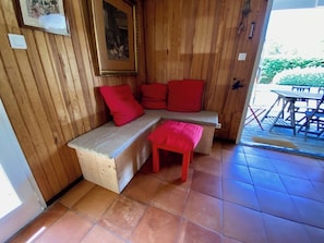 A wooden interior with a bench and a small stool, a framed picture on the wall, and a patio area visible through an open door. - Pool house in idyllic location A3695 (Lège-Cap-Ferret)