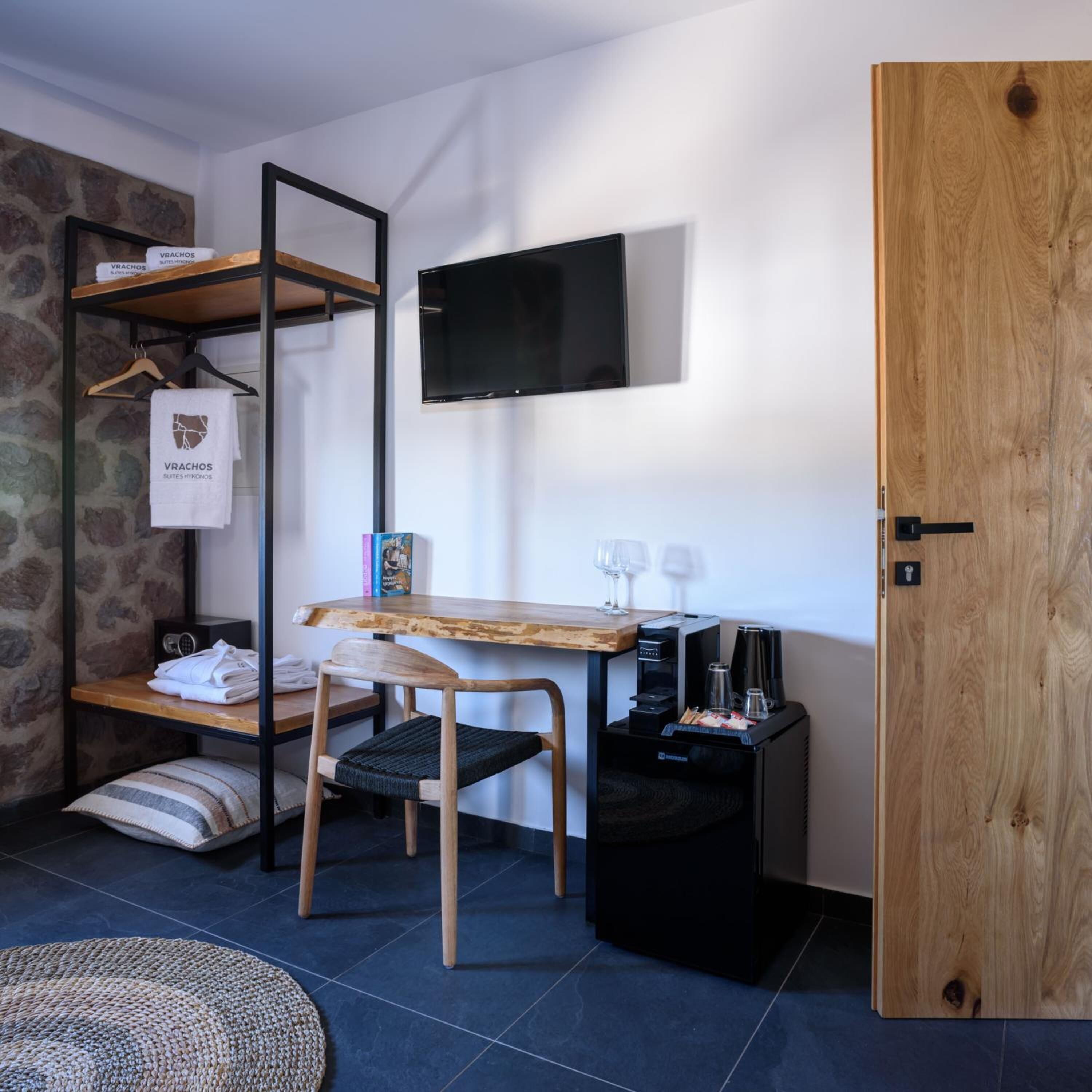 A compact room with a wooden desk, a chair, a TV mounted on the wall, a small black trash bin, and a wooden door.