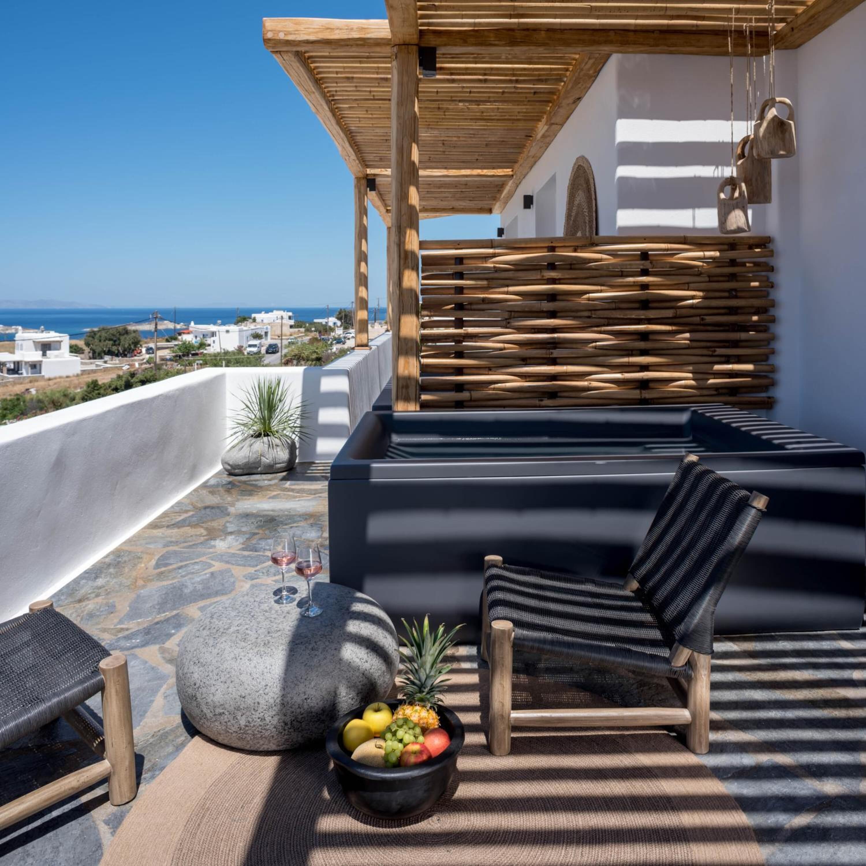Terraço no telhado com vista para o mar, apresentando área de estar com almofadas, parede de fibra e mesa de pedra com frutas.