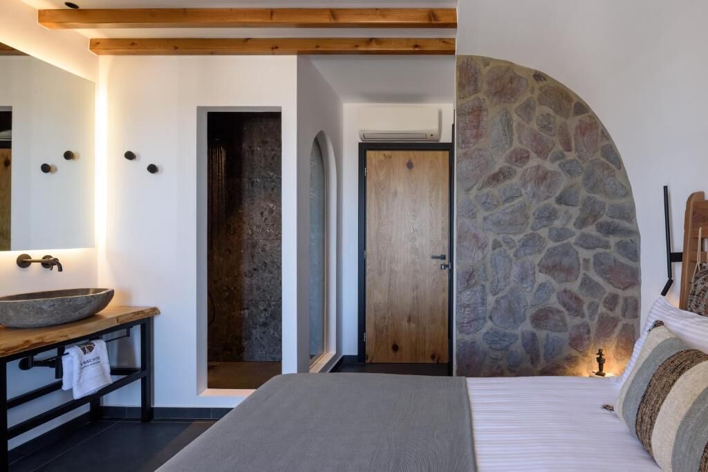 A bedroom with a stone wall, a wooden ceiling, a bed, a nightstand, a large round mirror, and a door leading to another room.