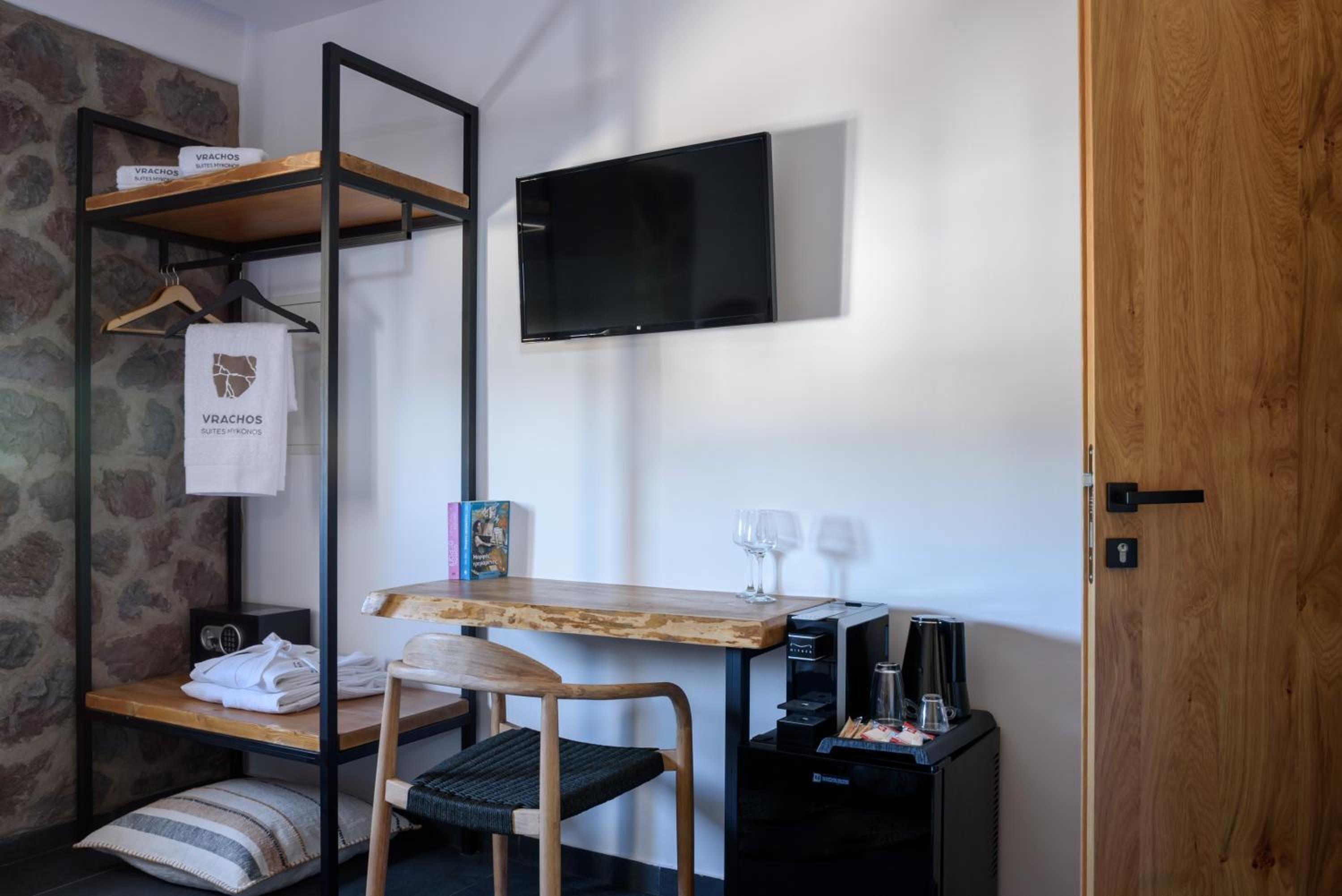 Une pièce de taille modeste, avec un bureau en bois, un téléviseur noir fixé au mur, une petite table avec des verres à vin, une chaise et une étagère contenant des serviettes et un vêtement suspendu.