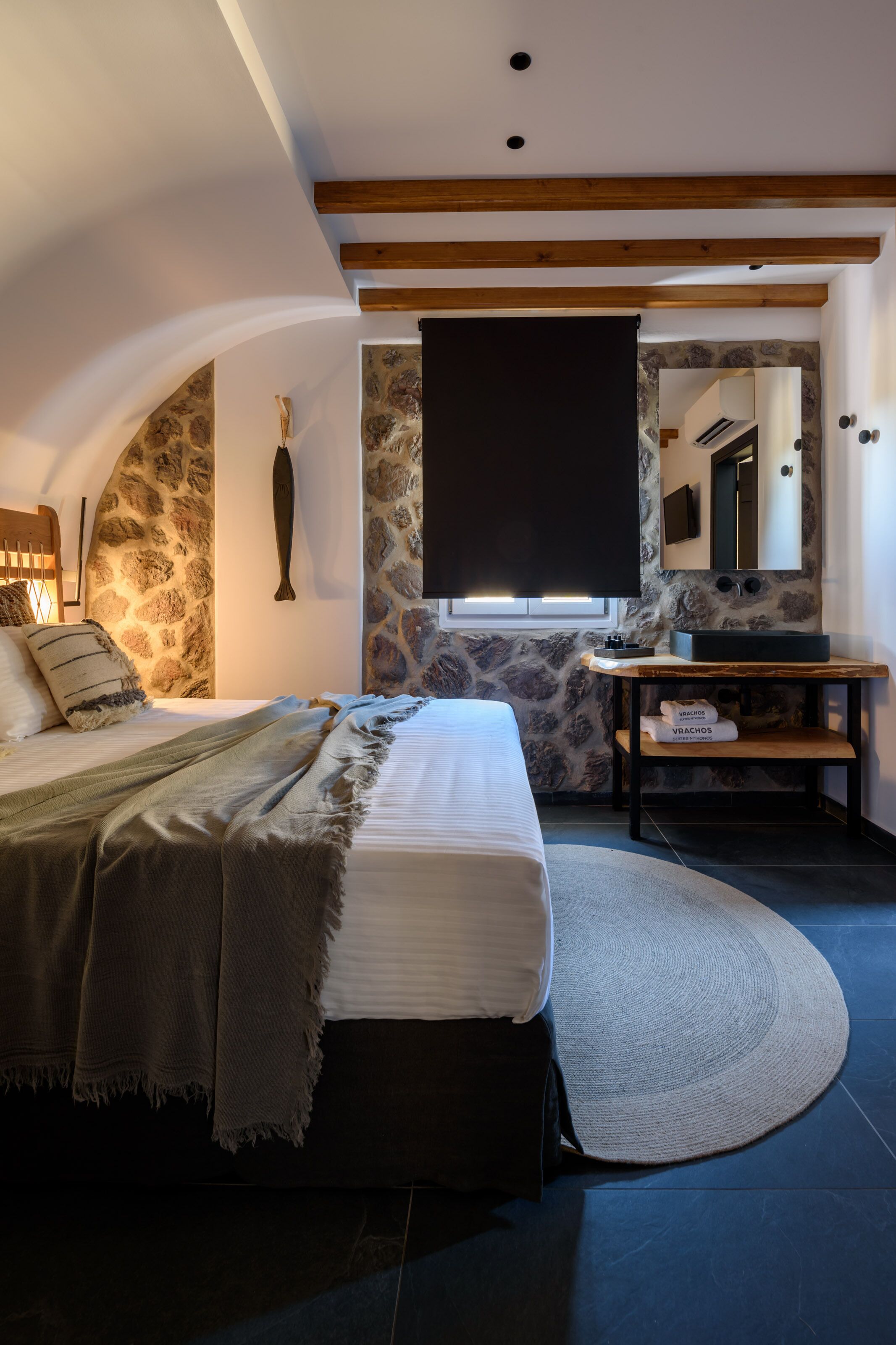 Un dormitorio con pared de piedra, una cama con colchón blanco y manta gris, cabecero de madera, una mesita de noche con espejo y un estante pequeño con toallas.