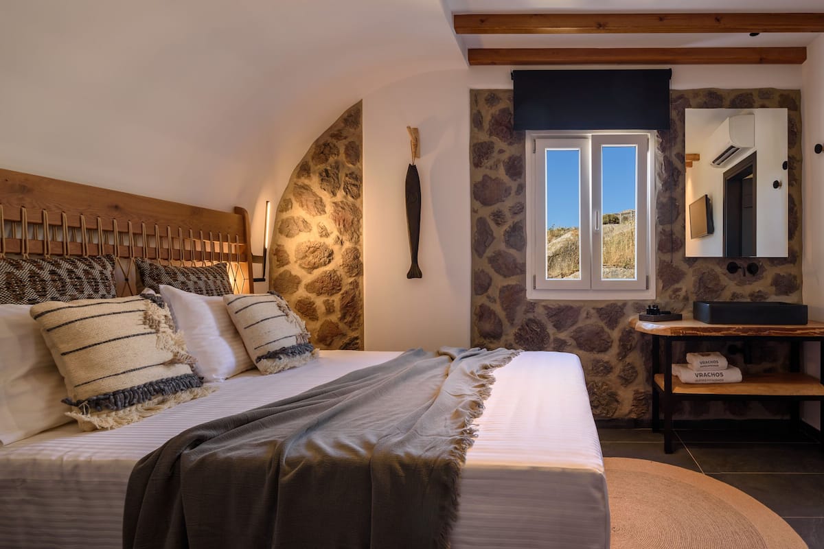 Un dormitorio con cama, pared de piedra, ventana, tocador con espejo y zona de ducha.