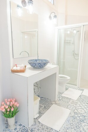 Un baño con un lavamanos blanco, un cuenco azul y blanco con estampado, un box de ducha de vidrio y un inodoro blanco.