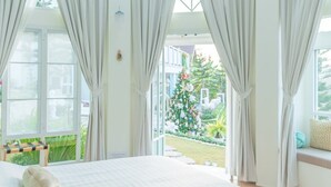 Un dormitorio con una cama grande, cortinas blancas, una araña y vistas a un árbol de Navidad decorado afuera.