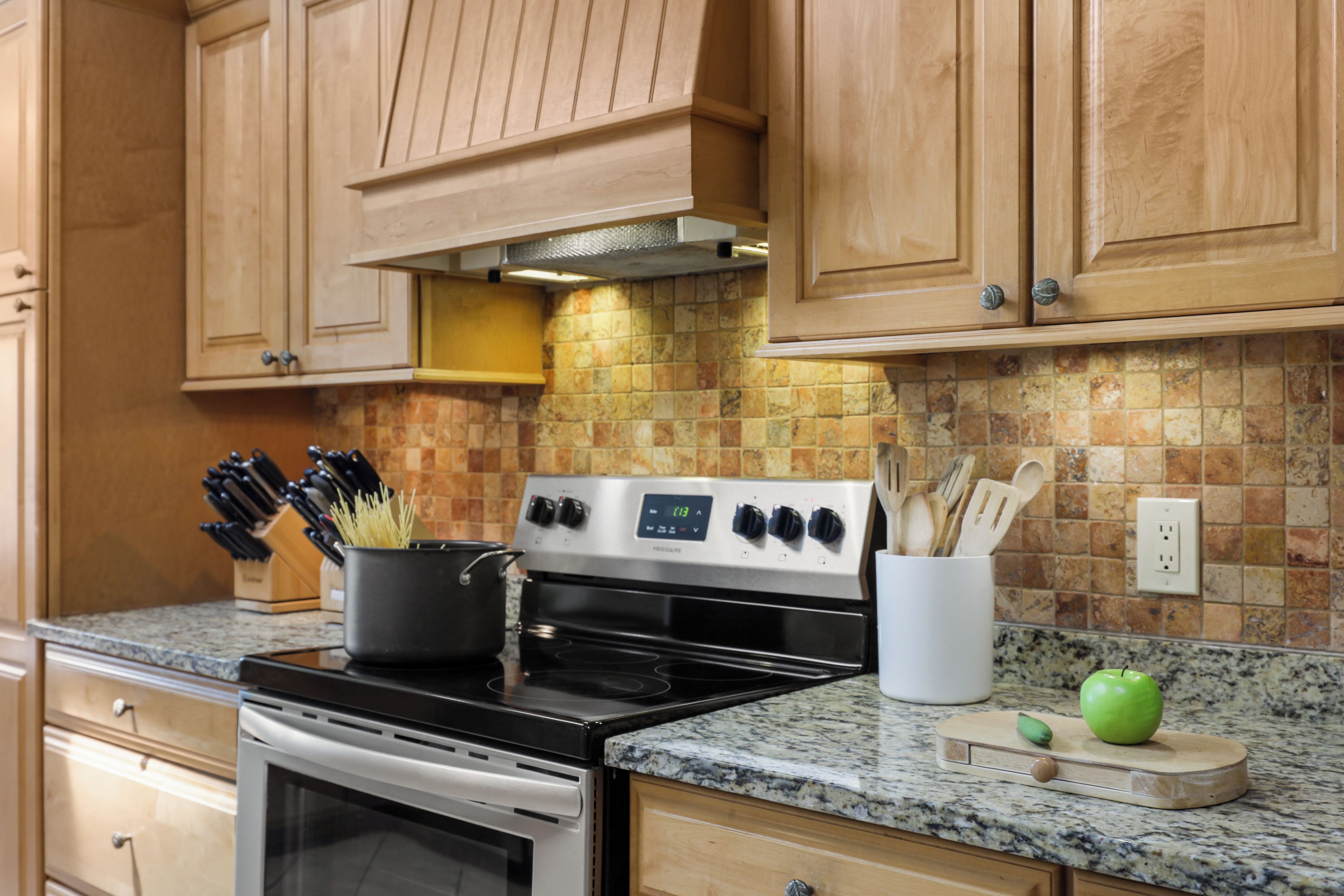 A kitchen with wooden cabinets, a stove, and a countertop with a knife block and a cutting board with an apple.