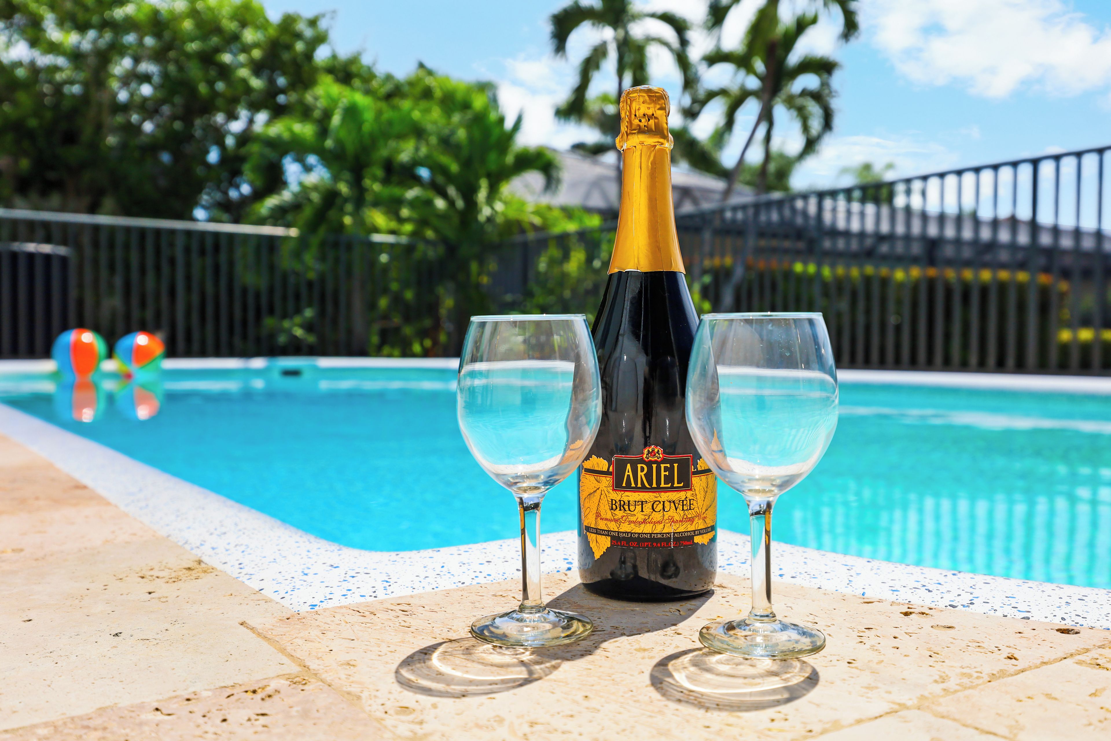 Een fles Ariel Brut Cuvée champagne en twee glazen bij een zwembad, met een helder blauw zwembad en een hek op de achtergrond.