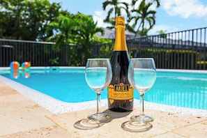 A bottle of Ariel Brut Cuvée champagne and two glasses on a poolside with a clear blue pool and a fence in the background.