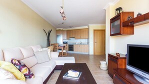 A modern living room with a sofa, coffee table, and TV stand. A kitchen with wooden cabinets and a dining area is visible in the background. - Home2Book La Tejita Beach and Pool (Granadilla)