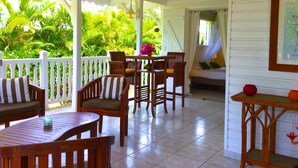 A covered patio with wooden furniture, a table, and chairs. There is a white railing and a view of lush greenery.