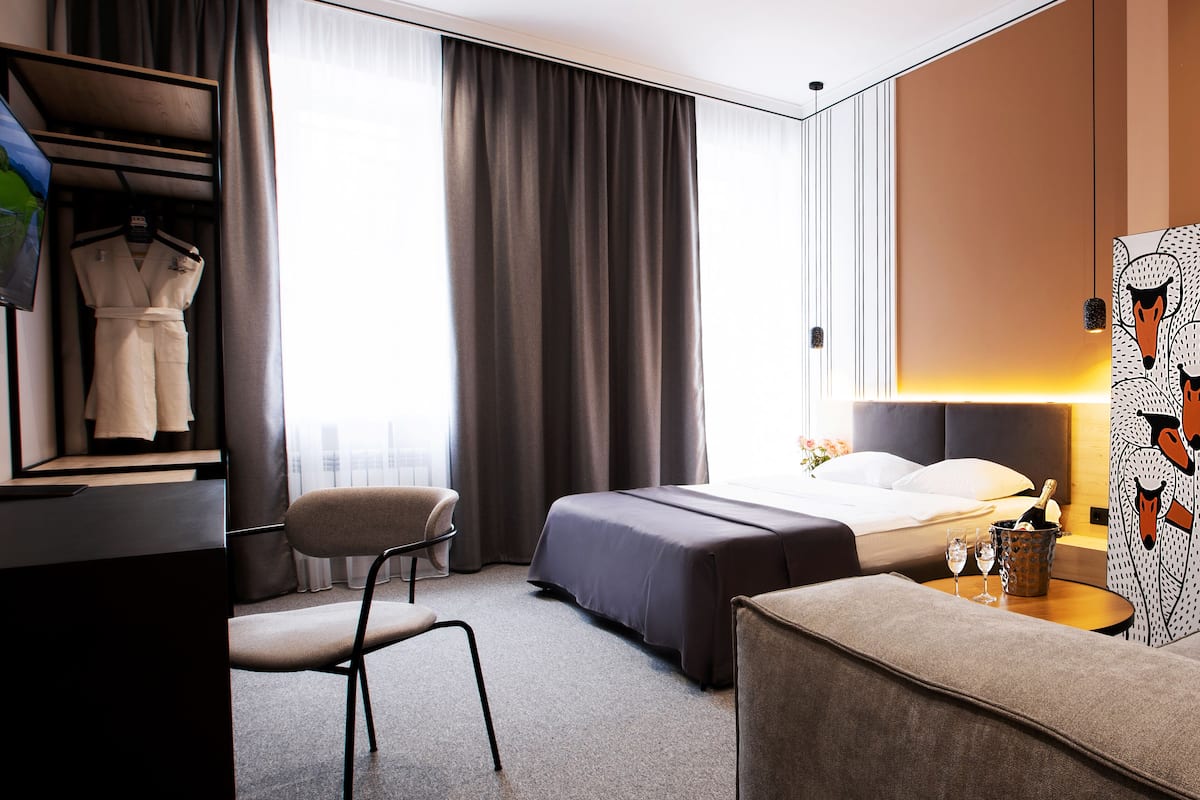 Une chambre d’hôtel moderne comprenant un lit, un bureau avec une chaise, une petite table avec une bouteille et des verres, et une grande fenêtre avec des rideaux.