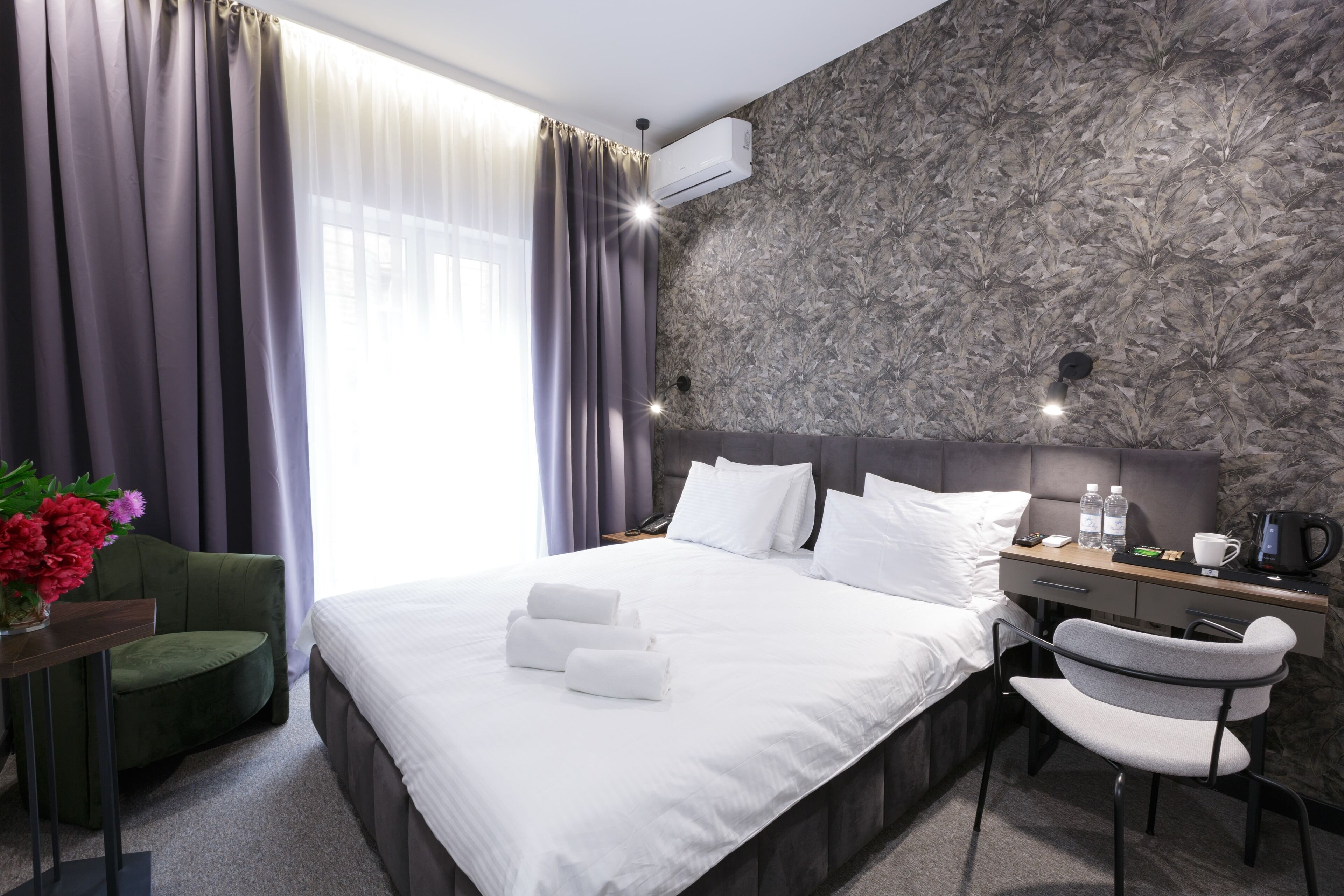 A modern hotel room with a large bed, a desk with a chair, a small table with a vase of flowers, and a window with curtains.