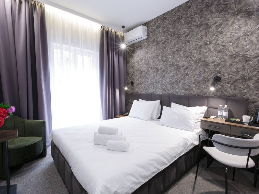 A modern hotel room with a large bed, a desk with a chair, a small table with a vase of flowers, and a window with curtains.