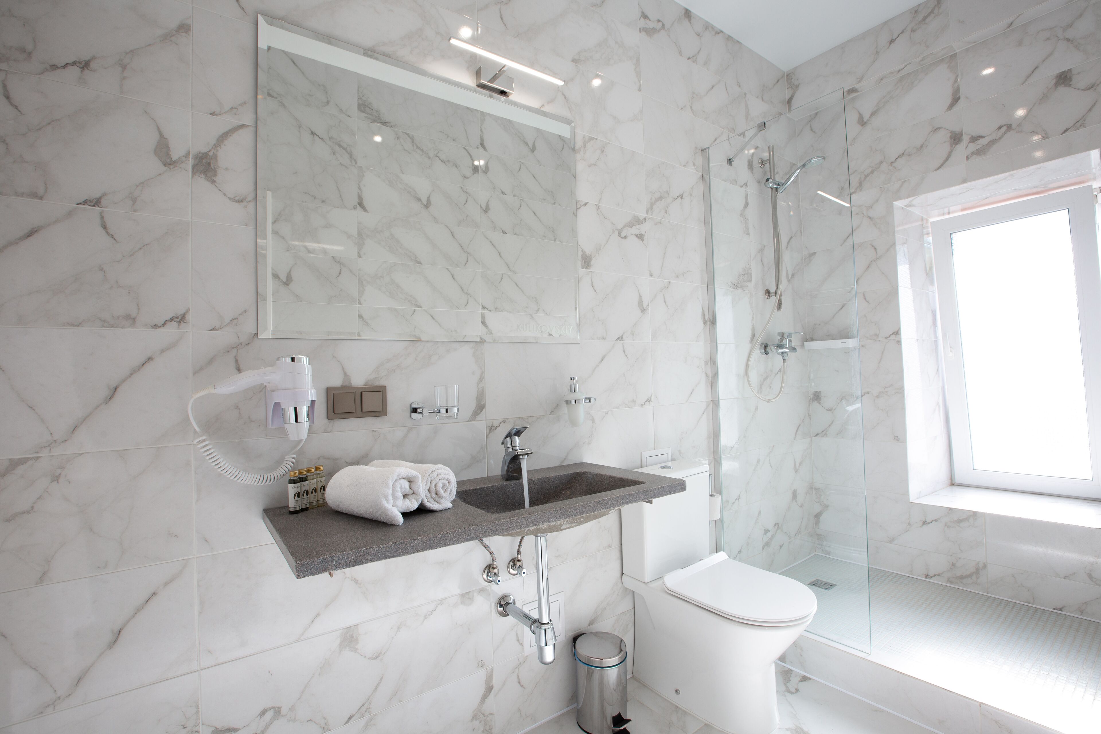 A modern bathroom with a marble wall, a glass shower enclosure, a white toilet, and a vanity with a sink and mirror.
