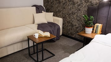 A hotel room with a sofa, a small wooden table with a book and a cup, a potted plant, and a stack of books on a nightstand.
