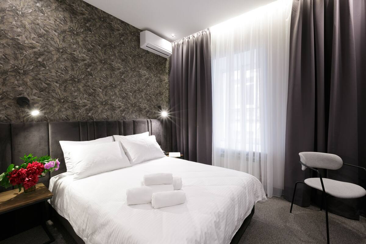 Une chambre d’hôtel moderne avec un grand lit, une tête de lit, une table de chevet ornée d’un vase de fleurs, une chaise et une fenêtre avec des rideaux.