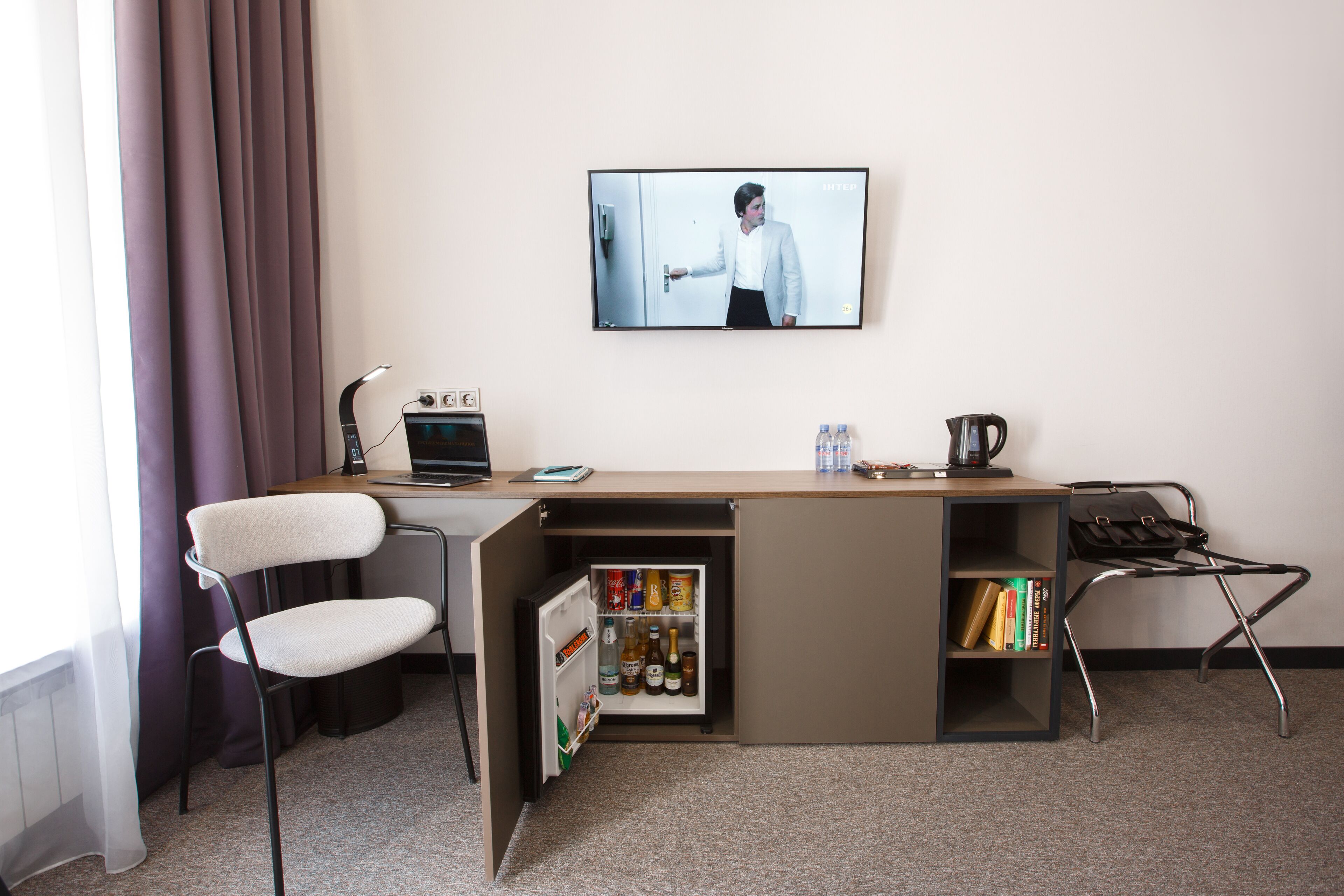 A compact hotel room with a kitchenette, a desk, a chair, a TV, and a bookshelf.