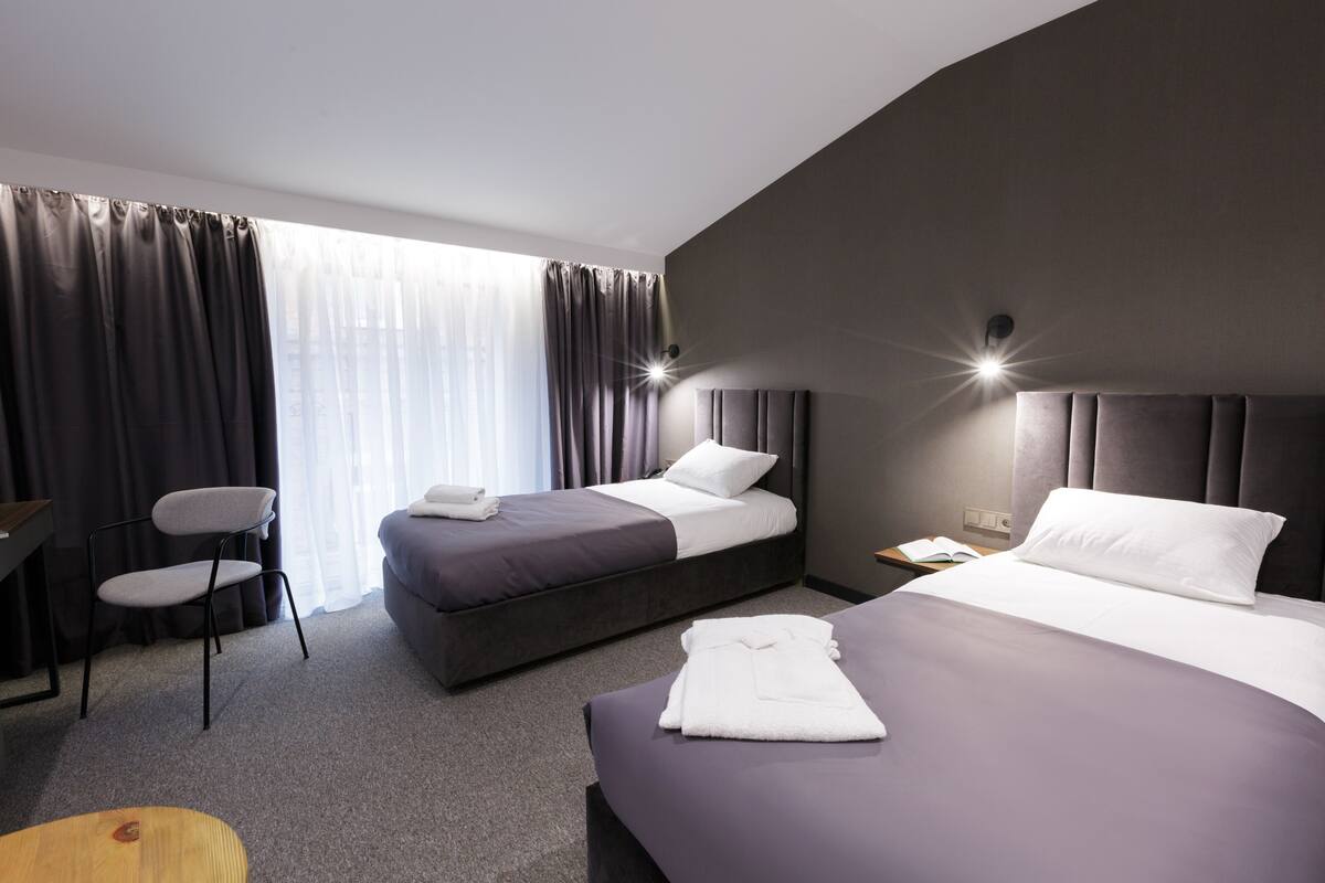 Une chambre d’hôtel avec deux lits, une chaise, un bureau et une fenêtre avec des rideaux.
