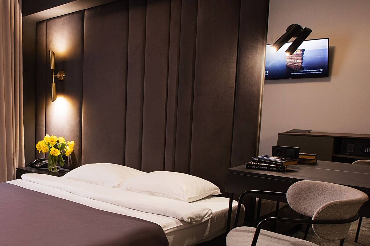 Une chambre d’hôtel avec un lit, un bureau, une chaise, un téléviseur fixé au mur et une table de chevet ornée d’un vase de fleurs.
