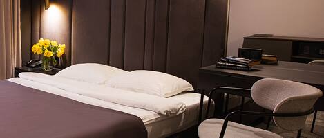 Una habitación de hotel con una cama, un escritorio, una silla, un televisor en la pared y una mesita de noche con un florero.